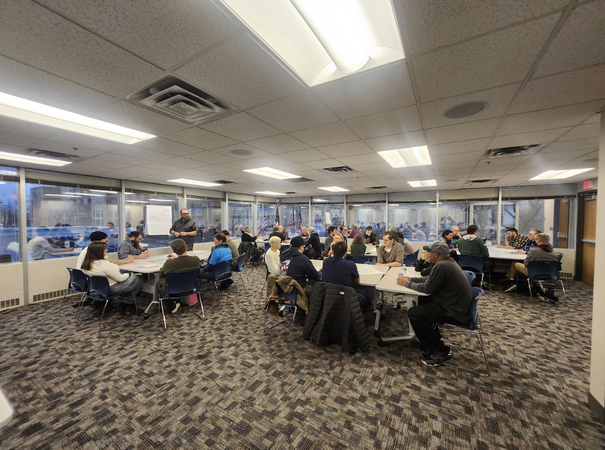 A room contains five tables, around each of which a small group is meeting