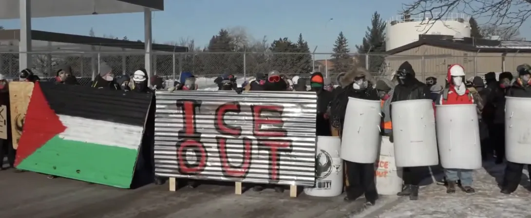 A group of masked people bundled up for winter, holding shields and two metal sheets that say "ICE OUT" and have the Palestinian flag